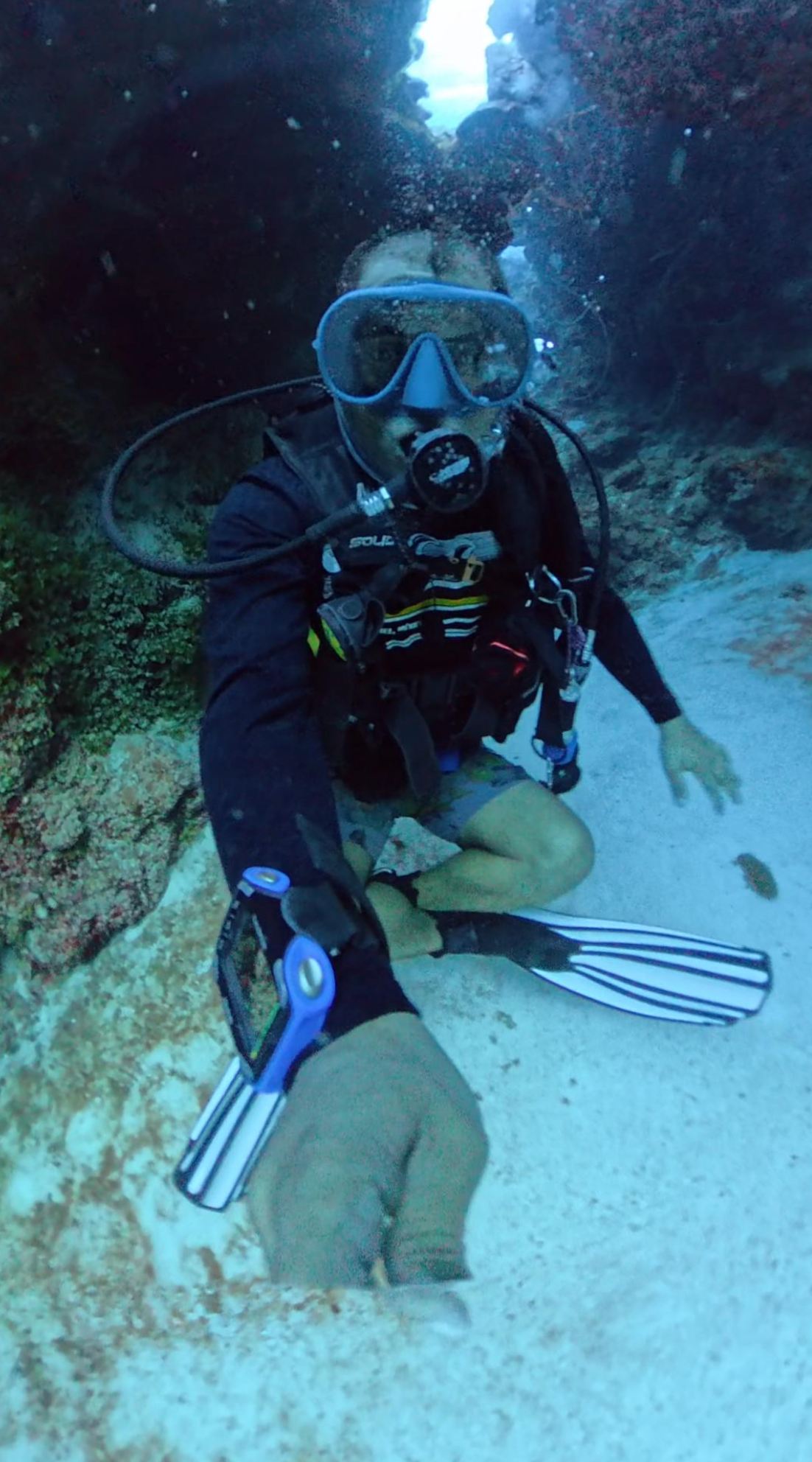A scuba diver wearing a mask, regulator, fins, and wetsuit kneels on a sandy ocean floor while taking a selfie underwater.